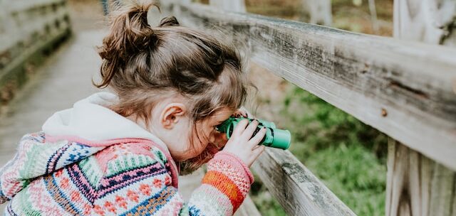 Child outdoors on wooden bridge holding binoculars to eyes